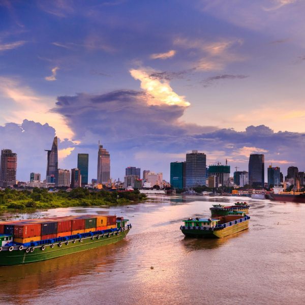 Container ships navigate the scenic Saigon River with Ho Chi Minh City skyline at dusk.